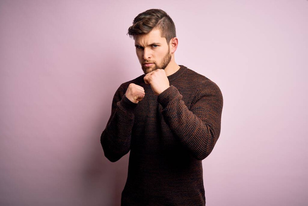 Young blond man with beard and blue eyes wearing casual sweater over pink background Ready to fight with fist defense gesture, angry and upset face, afraid of problem - Foto, imagen