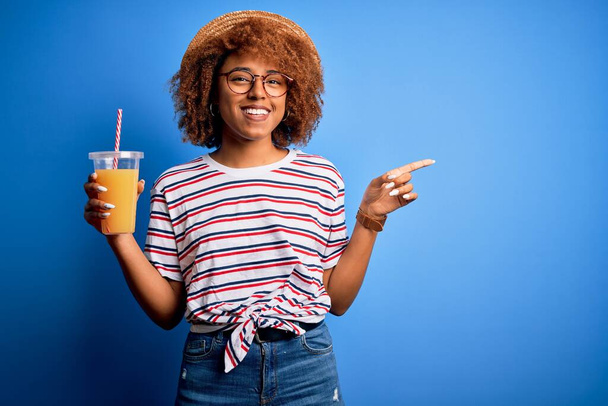 Młoda Afroamerykanka z kręconymi włosami na wakacjach w kapeluszu pijąca sok pomarańczowy bardzo szczęśliwa wskazując ręką i palcem z boku - Zdjęcie, obraz