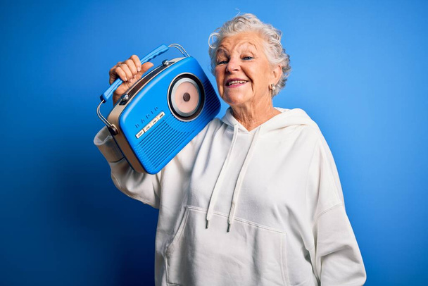Senior krásná žena drží vintage rádio stojící nad izolované modré pozadí s veselou tváří stojí a usmívá se s sebevědomým úsměvem ukazující zuby - Fotografie, Obrázek