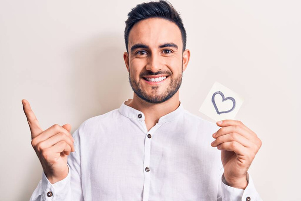 Jeune homme romantique beau avec barbe tenant rappel avec symbole de forme de coeur souriant heureux pointant avec la main et le doigt sur le côté - Photo, image