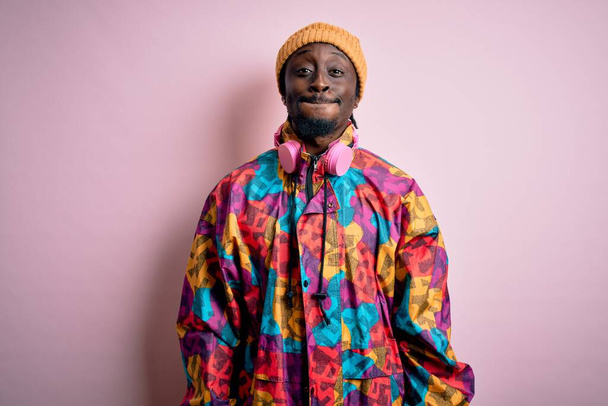 Junger gutaussehender afrikanisch-amerikanischer Mann in buntem Mantel und Mütze über rosa Hintergrund, der Wangen mit lustigem Gesicht aufbläst. Mund mit Luft aufgeblasen, verrückter Ausdruck. - Foto, Bild
