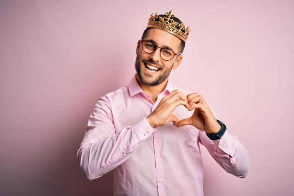 ピンクの背景の上に王として黄金の冠を身に着けている若いハンサムなビジネスマンは、手で心のシンボルと形状を示す愛に笑みを浮かべて。ロマン主義. - 写真・画像