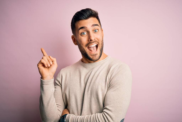 Junger gutaussehender Mann in lässigem Pullover vor isoliertem rosa Hintergrund mit einem breiten Lächeln im Gesicht, der mit Hand und Finger zur Seite zeigt und in die Kamera blickt. - Foto, Bild