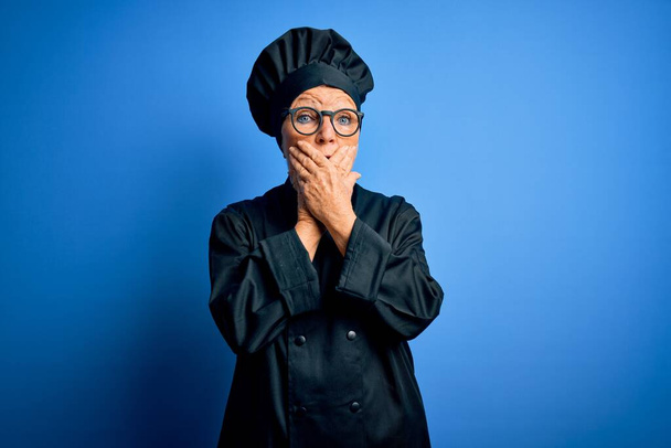 Senior beautiful grey-haired chef woman wearing cooker uniform and hat over blue background shocked covering mouth with hands for mistake. Secret concept. - Valokuva, kuva