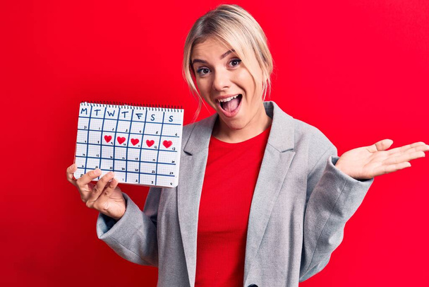 Блондинка, контролирующая календарь менструального периода на красном фоне, празднует достижение со счастливой улыбкой и выражением лица победителя с поднятой рукой - Фото, изображение
