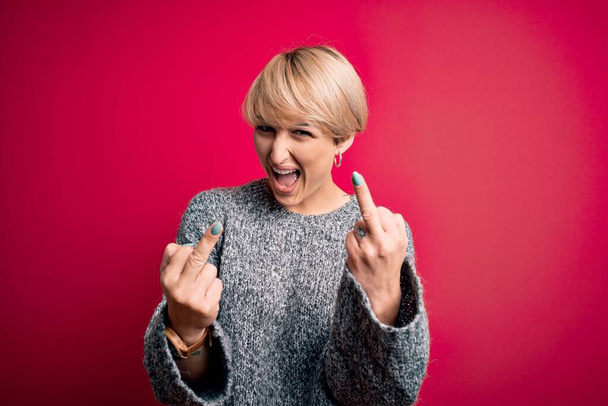 Young blonde woman with modern short hair wearing casual sweater over pink background Showing middle finger doing fuck you bad expression, provocation and rude attitude. Screaming excited - Foto, afbeelding