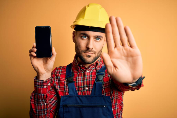 Fiatal építész férfi visel biztonsági sisak mutatja okostelefon képernyő sárga háttér nyitott kézzel csinál stop tábla komoly és magabiztos kifejezés, védelmi gesztus - Fotó, kép