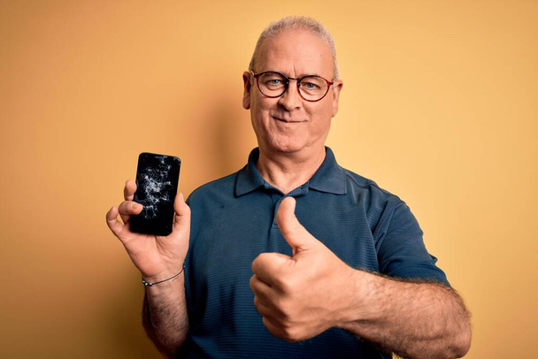 Middle age man holding broken smartphone showing cracked screen over yellow background happy with big smile doing ok sign, thumb up with fingers, excellent sign - Foto, Imagen