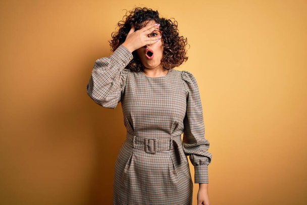 Beautiful arab business woman wearing dress and glasses standing over yellow background peeking in shock covering face and eyes with hand, looking through fingers with embarrassed expression. - Photo, Image