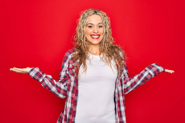 Mladá krásná blondýna žena na sobě ležérní košili stojící nad izolovaným červeným pozadím s úsměvem ukazující obě ruce otevřené dlaně, prezentační a reklamní srovnání a rovnováhu - Fotografie, Obrázek