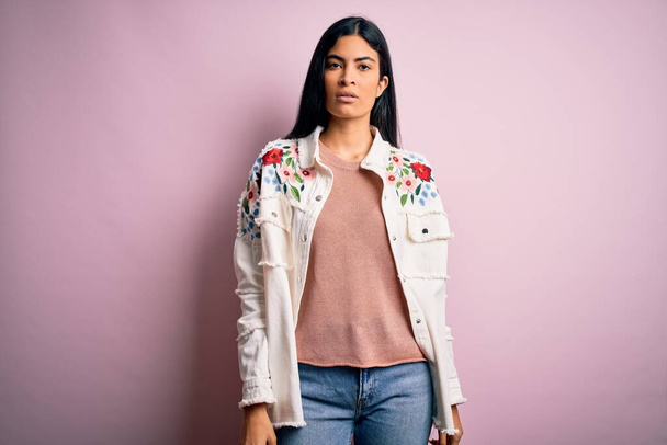 Young beautiful hispanic fashion woman wearing casual sweater over pink background with serious expression on face. Simple and natural looking at the camera. - 写真・画像