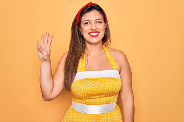 Junge hispanische Pin-up-Frau trägt Mode sexy 50er Jahre Stil auf gelbem Hintergrund zeigt und zeigt mit Finger Nummer vier, während sie selbstbewusst und glücklich lächelt. - Foto, Bild