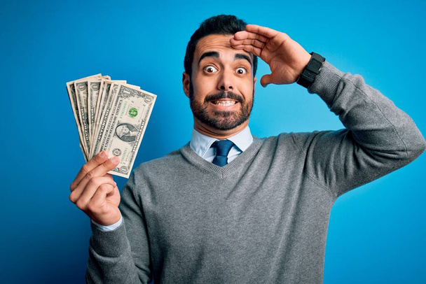 Young handsome man with beard holding bunch of dollars banknotes over blue background stressed with hand on head, shocked with shame and surprise face, angry and frustrated. Fear and upset for mistake. - Photo, Image