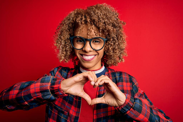 Jonge mooie Afrikaanse Amerikaanse vrouw draagt casual shirt en bril over rode achtergrond glimlachen in liefde tonen hart symbool en vorm met de handen. Romantisch concept. - Foto, afbeelding