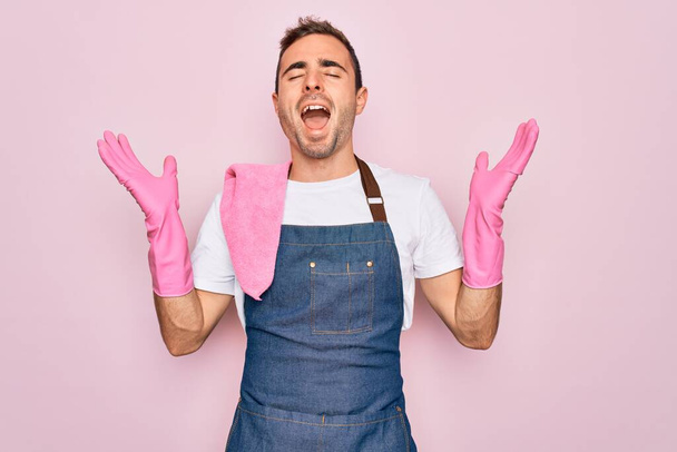 Νεαρός καθαριστής με μπλε μάτια που καθαρίζει φορώντας ποδιά και γάντια πάνω από ροζ φόντο γιορτάζει τρελός και τρελός για την επιτυχία με τα χέρια ψηλά και κλειστά μάτια ουρλιάζοντας ενθουσιασμένος. Έννοια νικητή - Φωτογραφία, εικόνα