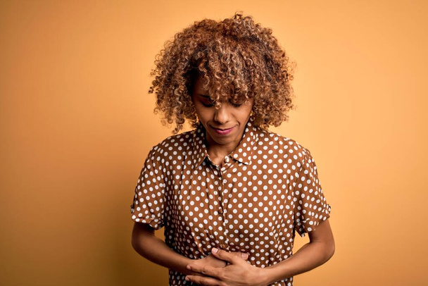 Junge schöne afrikanisch-amerikanische Frau in lässigem Hemd vor gelbem Hintergrund mit der Hand auf dem Bauch, weil Verdauungsstörungen, schmerzhafte Krankheit fühlen sich unwohl. Ache-Konzept. - Foto, Bild