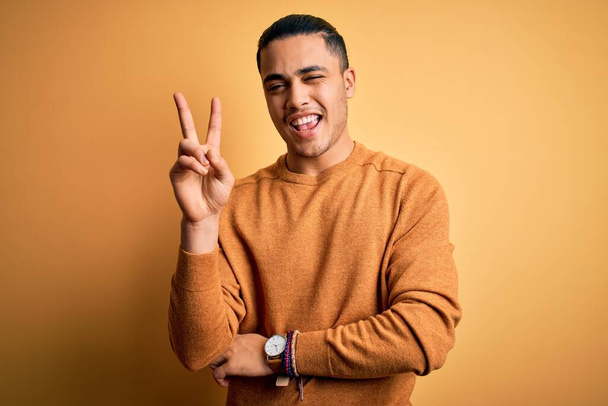 Young brazilian man wearing casual sweater standing over isolated yellow background smiling with happy face winking at the camera doing victory sign with fingers. Number two. - Photo, Image
