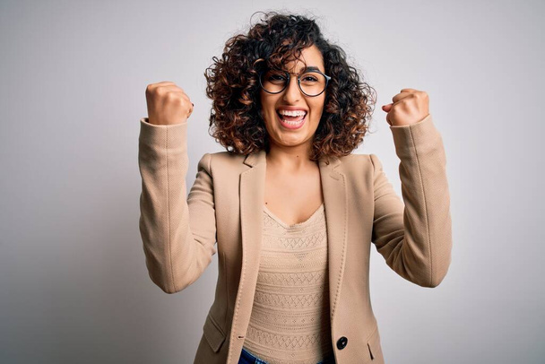Young curly arab business woman wearing elegant jacket and glasses over white background celebrating surprised and amazed for success with arms raised and open eyes. Winner concept. - Photo, Image