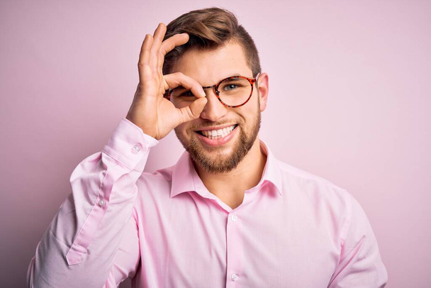 Jeune homme blond beau avec une barbe et des yeux bleus portant une chemise rose et des lunettes faisant un geste correct avec un sourire à la main, les yeux regardant à travers les doigts avec un visage heureux
. - Photo, image