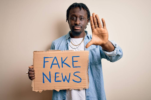 Jeune homme afro-américain tenant une bannière avec un faux message d'information sur fond blanc avec la main ouverte faisant stop signe avec expression sérieuse et confiante, geste de défense
 - Photo, image