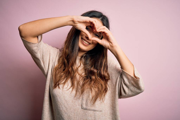 Young beautiful brunette woman wearing casual sweater standing over pink background Doing heart shape with hand and fingers smiling looking through sign - Photo, Image