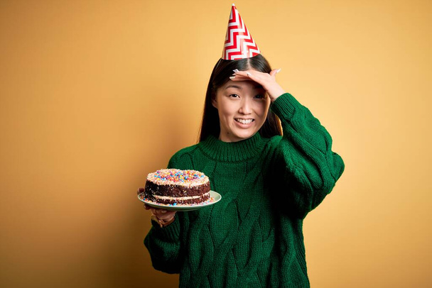 Young asian woman holding birthday cake wearing party hat over yellow isolated background stressed with hand on head, shocked with shame and surprise face, angry and frustrated. Fear and upset for mistake. - Photo, Image