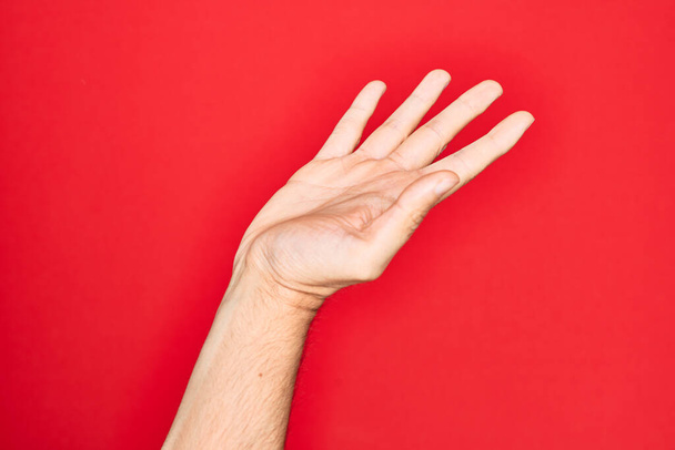 Hand of caucasian young man showing fingers over isolated red background presenting with open palm, reaching for support and help, assistance gesture - Valokuva, kuva