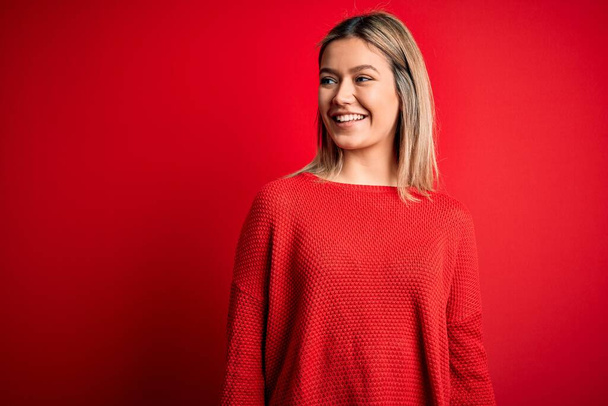 Young beautiful blonde woman wearing casual sweater over red isolated background looking away to side with smile on face, natural expression. Laughing confident. - Photo, Image
