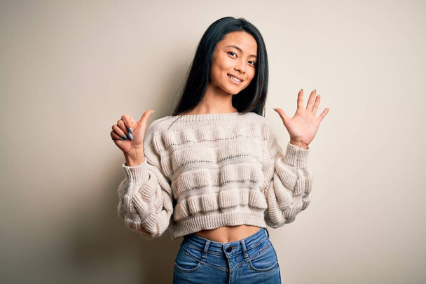 Young beautiful chinese woman wearing casual sweater over isolated white background showing and pointing up with fingers number six while smiling confident and happy. - Фото, изображение