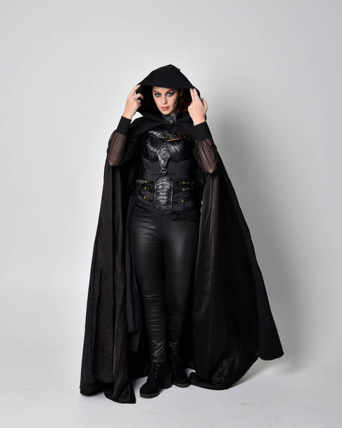 fantasy portrait of a woman with red hair wearing dark leather assassin costume with long black cloak. Full length standing pose  isolated against a studio background. - Valokuva, kuva