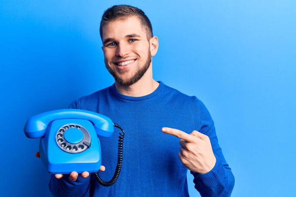 Joven hombre guapo sosteniendo teléfono vintage sonriendo feliz señalando con la mano y el dedo  - Foto, imagen
