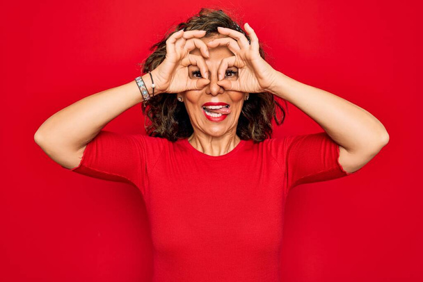 Mujer morena de mediana edad con una camiseta casual de pie sobre fondo rojo haciendo un buen gesto como binoculares sacando la lengua, ojos mirando a través de los dedos. Expresión loca
. - Foto, Imagen