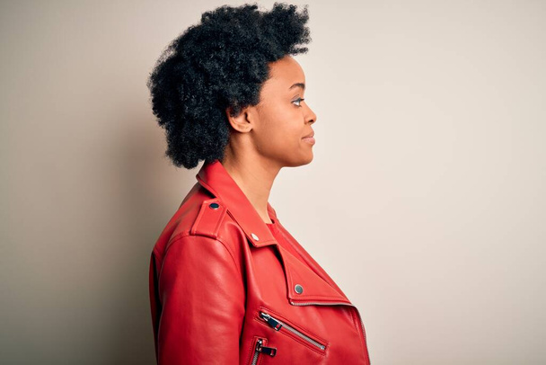 Jeune belle afro afro-américaine aux cheveux bouclés portant une veste rouge décontractée regardant vers le côté, pose de profil relax avec un visage naturel avec un sourire confiant
. - Photo, image
