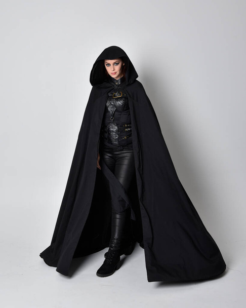 fantasy portrét ženy s rudými vlasy v tmavém koženém atentátnickém kostýmu s dlouhým černým pláštěm. Plná délka stojící póza izolované na pozadí studia. - Fotografie, Obrázek