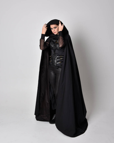 fantasy portrait of a woman with red hair wearing dark leather assassin costume with long black cloak. Full length standing pose  isolated against a studio background. - Φωτογραφία, εικόνα