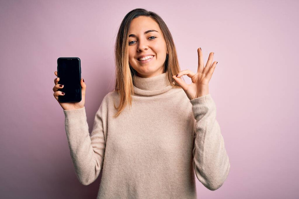 Giovane bella donna bruna in possesso di smartphone che mostra lo schermo su sfondo rosa facendo ok segno con le dita, simbolo eccellente
 - Foto, immagini