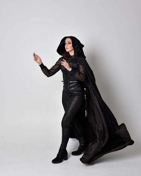 fantasy portrét ženy s rudými vlasy v tmavém koženém atentátnickém kostýmu s dlouhým černým pláštěm. Plná délka stojící póza izolované na pozadí studia. - Fotografie, Obrázek