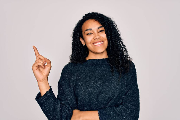 Молодая африканская кудрявая американка в обычном свитере стоит на белом фоне с широкой улыбкой на лице, указывая рукой и пальцем в сторону, глядя в камеру
. - Фото, изображение
