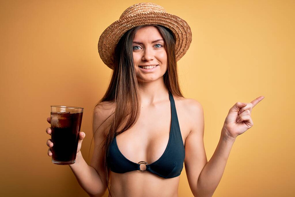 Joven hermosa mujer con ojos azules en vacaciones con bikini y sombrero bebiendo bebida cola muy feliz señalando con la mano y el dedo a un lado
 - Foto, Imagen