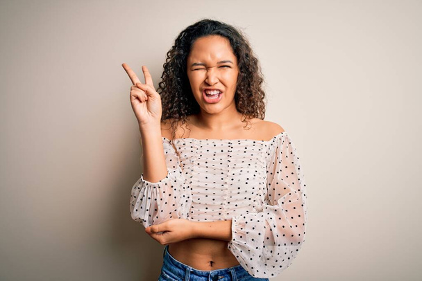 Young beautiful woman with curly hair wearing casual t-shirt standing over white background smiling with happy face winking at the camera doing victory sign. Number two. - Фото, изображение