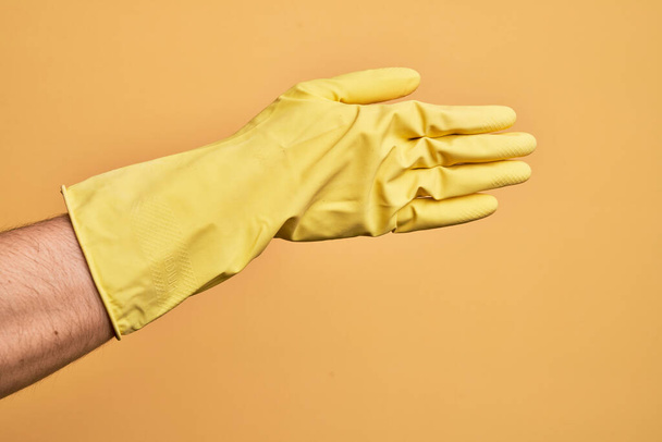 Χέρι καυκάσιου νεαρού άνδρα με γάντι καθαρισμού πάνω από απομονωμένο κίτρινο φόντο που τεντώνεται και φτάνει με ανοιχτό χέρι για χειραψία, δείχνοντας το πίσω μέρος του χεριού - Φωτογραφία, εικόνα