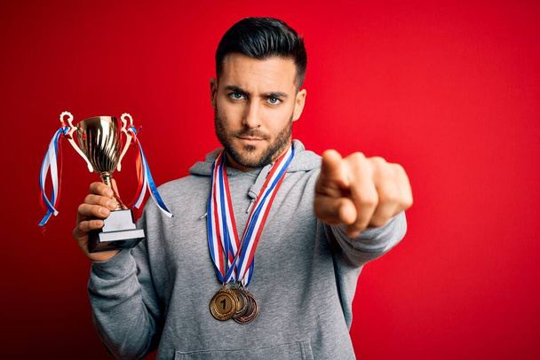 Jeune homme joyeux et victorieux tenant un trophée portant des médailles sur fond rouge pointant du doigt vers la caméra et vers vous, signe de la main, geste positif et confiant de devant - Photo, image
