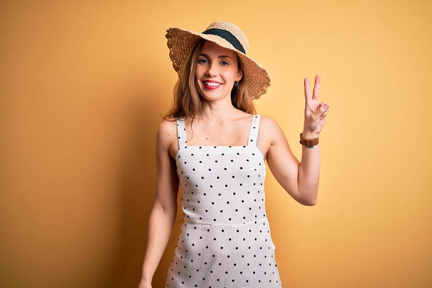 Mladá krásná blondýna na dovolené na sobě letní klobouk přes žluté pozadí s úsměvem šťastný obličej mrkající na kameru dělat vítězství znamení. Číslo dvě. - Fotografie, Obrázek