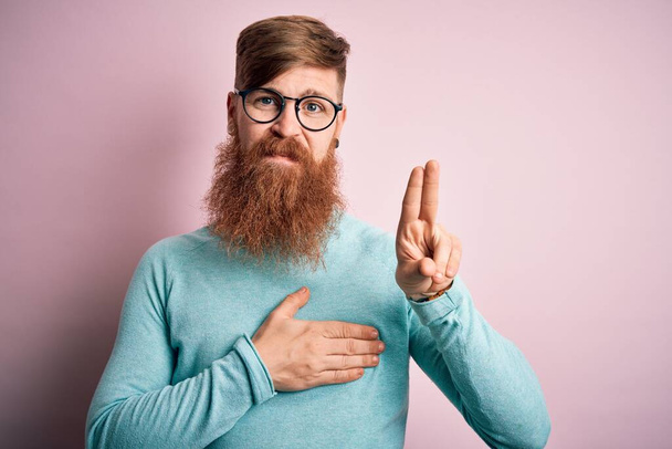 Hezký irský rusovous muž s vousy nosí brýle přes růžové izolované pozadí s úsměvem s rukou na hrudi a prsty nahoru, dělat věrnostní slib - Fotografie, Obrázek