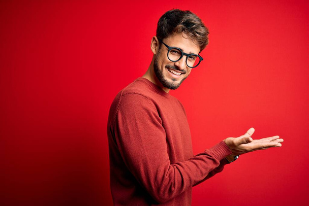 Young handsome man with beard wearing glasses and sweater standing over red background pointing aside with hands open palms showing copy space, presenting advertisement smiling excited happy - Photo, image