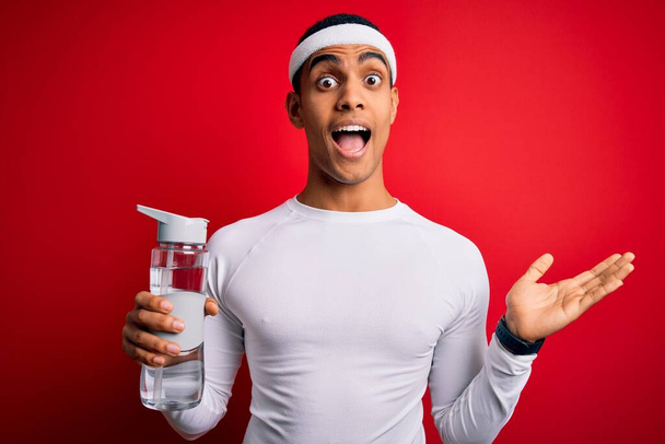Young handsome african american sportsman wearing sportswear drinking bottle of water very happy and excited, winner expression celebrating victory screaming with big smile and raised hands - Fotó, kép