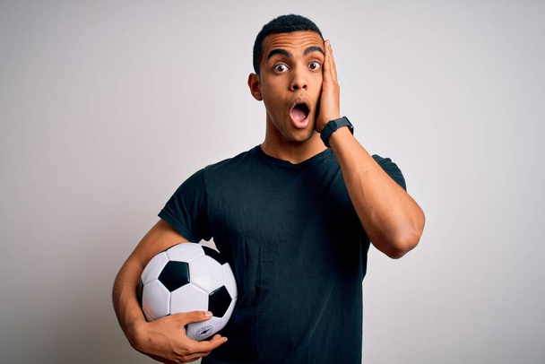 Przystojny Afroamerykanin gra w piłkę nożną trzymając piłkę na białym tle boi się i zszokowany, zaskoczony i zdumiony wyraz twarzy z rękami na twarzy - Zdjęcie, obraz