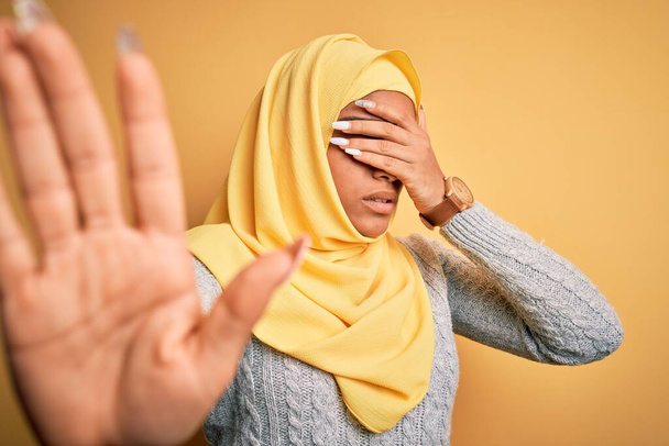 Young beautiful african american girl wearing muslim hijab over isolated yellow background covering eyes with hands and doing stop gesture with sad and fear expression. Embarrassed and negative concept. - Valokuva, kuva