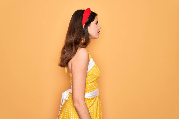 Молодая красивая пин-ап женщина носить 50-х годов моды винтажное платье на желтом фоне глядя в сторону, расслабиться профиль позировать с естественным лицом с уверенной улыбкой
. - Фото, изображение
