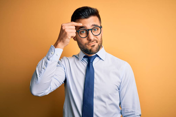 Young handsome businessman wearing tie and glasses standing over yellow background pointing unhappy to pimple on forehead, ugly infection of blackhead. Acne and skin problem - Foto, Imagen